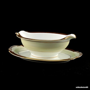 Puede incluir: Una salsera blanca con un platillo a juego. La salsera y el platillo tienen un ribete dorado y un acento marrón en el borde. La salsera tiene un asa curva y un pico vertedor.