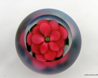 May include: A round glass paperweight featuring a vibrant red flower with green center. The flower is encased within a clear glass sphere, surrounded by a gradient of pink, blue, and black. The paperweight is a decorative item.