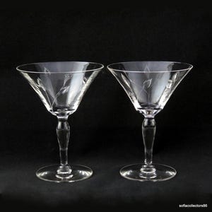 Könnte beinhalten: Zwei klare Martini-Gläser mit geätzten floralen Designs. Die Gläser haben eine klassische konische Schalenform, einen schlanken Stiel und einen kreisförmigen Fuß. Das geätzte Design zeigt zarte, stilisierte Blätter. Der Hintergrund ist schwarz.