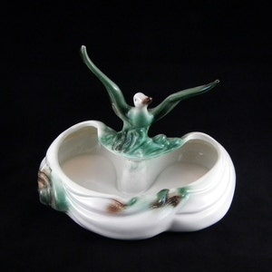 以下が含まれることがあります： 緑と茶色のアクセントが付いた白いセラミックプランター。翼を広げた様式化された鳥が特徴です。鳥はプランターの2つの開いたコンパートメントの上に配置され、装飾的で機能的な作品を作り出しています。