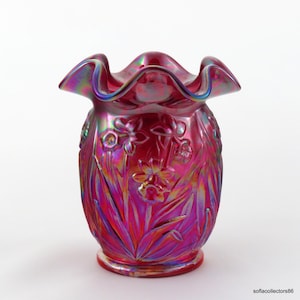 May include: A vibrant, iridescent pink vase with a ruffled rim. The vase features a floral design with daffodils and leaves. The glass has a glossy finish, reflecting a rainbow of colors. The base is a solid pink.