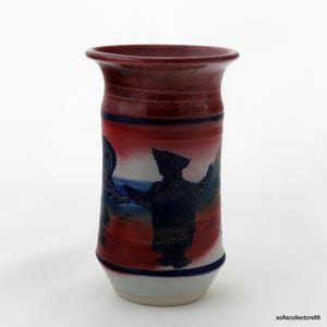 May include: A handmade ceramic vase with a flared rim. The vase features a red and blue color scheme with a painted silhouette of two figures. The base is white, and the vase is approximately 6 inches tall.