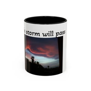 May include: White ceramic mug with a black interior and rim. The mug features a vibrant sunset scene with red and orange clouds over a silhouette of trees. The text "this storm will pass" is printed on the mug.