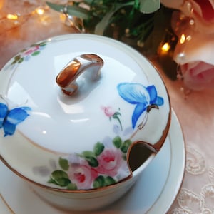 May include: A white porcelain serving dish with a lid, featuring hand-painted blue butterflies and pink roses. The dish has a gold trim and a gold-colored handle on the lid. It sits on a matching saucer, creating a classic, elegant look.