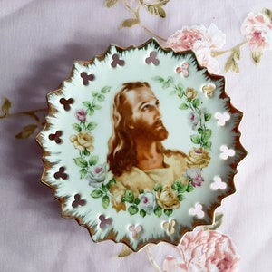 May include: A decorative white porcelain plate with a gold rim and a central image of Jesus. The plate is adorned with a floral wreath of pink, yellow, and white roses. The plate has a scalloped edge with clover-shaped cutouts.