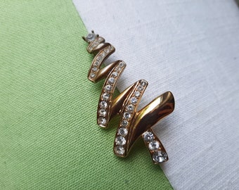 May include: Gold-coloured brooch in a zigzag design, set with clear rhinestones. The brooch has a polished finish and a pin fastening. The background is a green and white fabric.