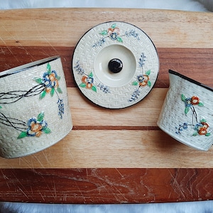 May include: A vintage tea set with a cream-colored basketweave design. The set includes a cup, a creamer, and a lid, all adorned with floral patterns in blue, orange, and green. The cup has a black handle and rim.