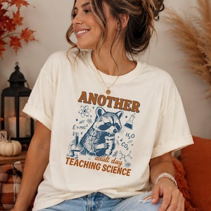 May include: A cream-colored short-sleeve t-shirt featuring a graphic of a raccoon surrounded by science-themed icons including an atom, a beaker, and chemical formulas. The text reads "ANOTHER adult day TEACHING SCIENCE" in brown.