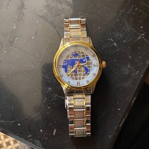 Puede incluir: Un reloj de pulsera dorado y plateado con una esfera blanca con un diseño de mapa azul. El reloj tiene manecillas e indicadores de hora dorados y una correa plateada y dorada. El reloj está sobre una superficie oscura.