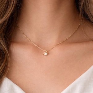 May include: A delicate gold necklace featuring a small, white pendant. The pendant has a smooth, rounded form, and the chain is fine and elegant. The necklace is worn against a white top.