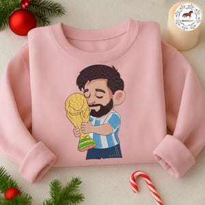 Puede incluir: Una sudadera de cuello redondo color rosa claro con una ilustraci&oacute;n bordada de estilo caricatura de una persona con barba y cabello oscuro, vistiendo una camiseta a rayas azul claro y blanca, abrazando un gran trofeo dorado con detalles verdes.