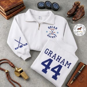 Op de afbeelding: Witte trui met een kwart rits met het "Briar University Hockey" logo en gekruiste hockeysticks op de mouw. Een tweede wit shirt toont "GRAHAM 44" in blauw. Hockeypucks, schaatsen en een fluitje zijn ook zichtbaar.