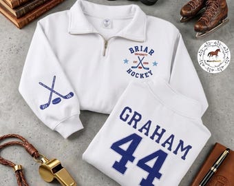 Op de afbeelding: Witte trui met een kwart rits met het "Briar University Hockey" logo en gekruiste hockeysticks op de mouw. Een tweede wit shirt toont "GRAHAM 44" in blauw. Hockeypucks, schaatsen en een fluitje zijn ook zichtbaar.