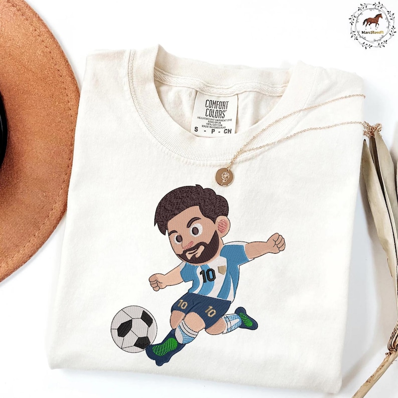 Puede incluir: Una camiseta de manga corta color blanco roto con un bordado de un jugador de f&uacute;tbol de dibujos animados con barba, que viste una camiseta a rayas azules y blancas con el n&uacute;mero 10, pantalones cortos azul oscuro y botas verdes, pateando un bal&oacute;n.