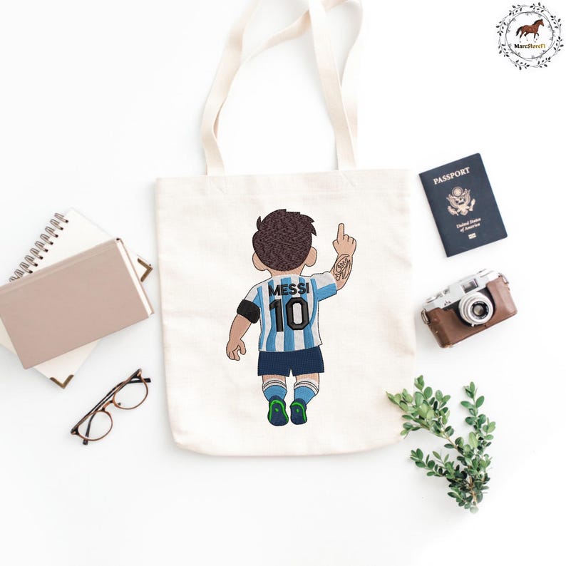 Puede incluir: Una bolsa de tela color crema con una ilustraci&oacute;n bordada de un jugador de f&uacute;tbol visto desde atr&aacute;s, que lleva una camiseta a rayas azul claro y blanca con el nombre MESSI y el n&uacute;mero 10, pantalones cortos oscuros y botas verdes y azules.
