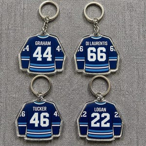 May include: Four acrylic keychains shaped like dark blue hockey jerseys with white stripes and numbers. Each keychain features a unique name and number: GRAHAM 44, DI LAURENTIS 66, TUCKER 46, and LOGAN 22. Each jersey is attached to a silver metal ring.
