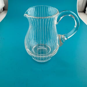 May include: A clear glass pitcher featuring a delicate white vertical pinstripe pattern throughout its rounded body. The vessel includes a curved handle on the side and a small pouring spout at the top rim, showcasing a transparent design.