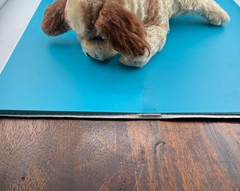 May include: A plush toy dog in a lying position with cream-colored fur and brown patches on its ears, head, and back. The stuffed animal features long, floppy ears, a small black nose, and a soft, textured exterior, captured from a high angle.
