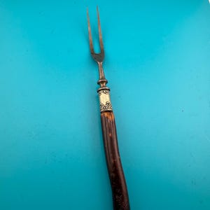 以下が含まれることがあります： ダークブラウンの質感のあるハンドルと、精巧なエンボス加工が施されたシルバー調の金属製フェルールを備えた2本歯のカービングフォーク。金属製の歯は細く、経年による摩耗や酸化の跡が見られます。