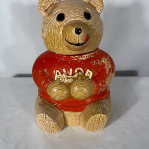 Puede incluir: Un tarro de galletas de cerámica con forma de oso y acabado en color canela texturizado, que lleva un suéter rojo con la palabra AVON en letras blancas en relieve. El oso tiene ojos negros, nariz negra y una pequeña lengua roja visible. La pintura roja del suéter muestra un desgaste y desconchado significativos en la parte frontal.