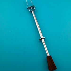 May include: A silver-toned metal kitchen tool featuring a two-pronged fork at one end and a dark brown wooden handle at the other. The tool includes a sliding mechanism with a circular metal disc along the central shaft for ejecting food items.
