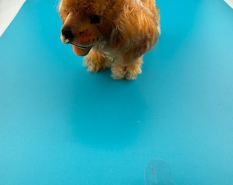 May include: A plush toy dog with golden-brown fur, long floppy ears, and a black nose. The stuffed animal features a small collar around its neck and is depicted in a standing position with a soft, textured coat and detailed facial features.
