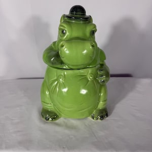 May include: A glossy lime green ceramic cookie jar shaped like a standing hippopotamus wearing a small black bowler hat. The hippo has a rounded belly, visible suspender buttons, and dark shading on the hat, feet, and facial features.