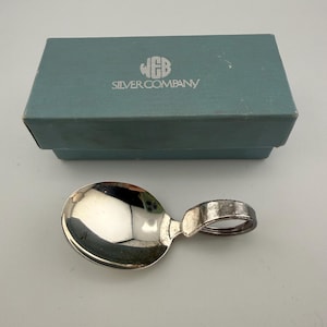 May include: A silver-colored spoon with a short, rounded handle loop positioned in front of a rectangular, light blue box. The box features the text WEB SILVER COMPANY printed in white on the lid, with a logo above the brand name.