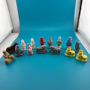 Op de afbeelding: Een verzameling kleine, gegoten beeldjes in diverse kleuren en vormen, waaronder sneeuwpoppen, kanonnen, figuren met hoge hoeden, dieren en abstracte vormen. Deze decoratieve miniatuuritems zijn in een losse rij geplaatst en vertonen uiteenlopende texturen en effen kleuren zoals rood, blauw, geel, wit en bruin.
