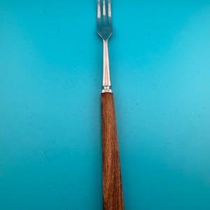 May include: A stainless steel fork featuring a long, slender, polished metal head with three tines and a straight, dark brown wooden handle with a visible grain pattern. The handle is cylindrical and tapers slightly toward the metal neck.