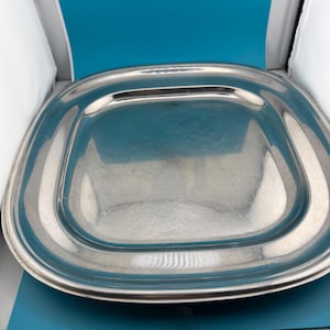 May include: A square stainless steel tray with rounded corners and a polished, reflective surface. The tray features a wide, flat rim that transitions into a shallow, recessed center area, showcasing a smooth, metallic finish throughout.