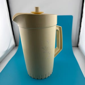 Può includere: Una caraffa in plastica color crema con superficie testurizzata e un sottile motivo floreale. È dotata di un manico robusto, un ampio beccuccio per versare e un coperchio abbinato con un piccolo pomello circolare giallo sulla parte superiore.