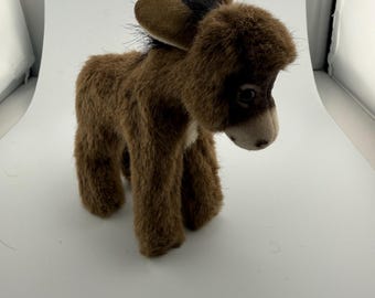 May include: A plush donkey toy with brown fur, a black mane, and a light-colored muzzle. The stuffed animal features large, upright ears, dark eyes, and a soft, textured coat. It stands on four legs and has a small metallic button on one ear.