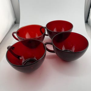 May include: A set of four deep red glass teacups with rounded bowls and small side handles. The cups are made of translucent, dark crimson glass, featuring a smooth, polished finish and a consistent circular shape across all four individual pieces.