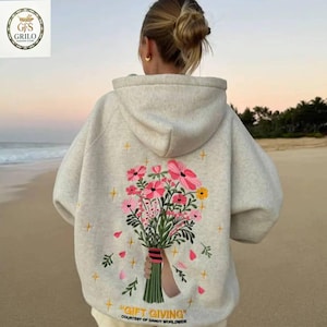 May include: A light gray hoodie with a floral embroidery design on the back. The embroidery features pink and yellow flowers, green stems, and the text "GIFT GIVING" at the bottom. The hoodie has a hood and long sleeves.