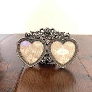 May include: A double heart-shaped picture frame made of antique silver-toned metal. The frame features a decorative floral motif with roses and leaves at the top and bottom. The two heart openings contain beige marbled paper inserts, with the Hallmark logo.