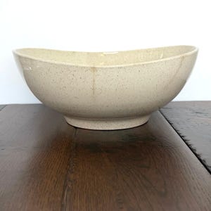 May include: A speckled, oval-shaped ceramic bowl in a neutral beige color. The bowl has a wide opening and a slightly raised base. The interior is a lighter shade of beige. The bowl is likely used for serving food or as a decorative piece.