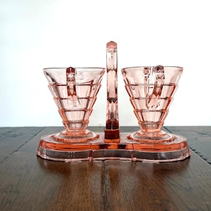 May include: A pink glass sugar and creamer set. The set includes two handled cups and a central handle, all connected by a rectangular base. The cups have a geometric design. The set is made of pink glass.