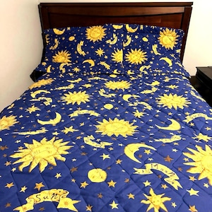 May include: A quilted bedspread and two matching pillow shams featuring a deep blue background with a repeating pattern of yellow suns, crescent moons, and stars. The design includes small banners with the printed words Sun and Moon.