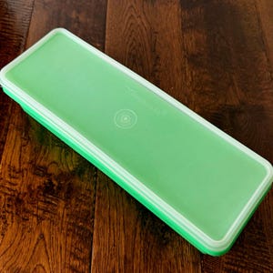 May include: A rectangular light green Tupperware storage container with a matching lid. The lid features a white border, a centered circular indentation, and the embossed brand name Tupperware clearly visible on the top surface.