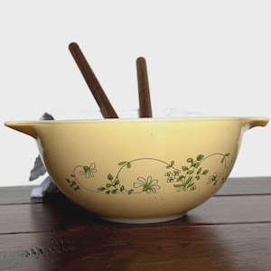 May include: A vintage yellow mixing bowl with a floral design in green. The bowl has a handle on each side and is resting on a wooden surface. Two wooden spoons are inside the bowl.