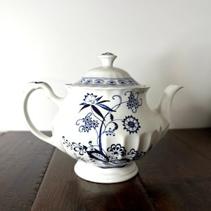 May include: A white ceramic teapot featuring a blue floral pattern with intricate botanical illustrations. The teapot has a rounded body with vertical fluted detailing, a matching lid with a small knob, a curved handle, and a tapered spout.