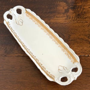 May include: An elongated white ceramic serving tray featuring a scalloped edge and gold-toned floral trim along the border. The tray includes two integrated handles at each end, each decorated with a raised shell-like motif and gold accents.