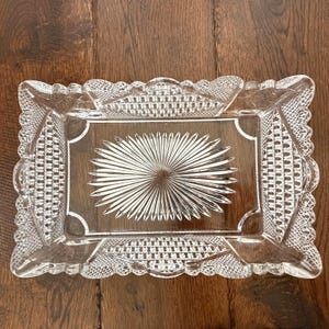 May include: Clear glass rectangular serving tray with a starburst design in the center. The tray has a decorative, textured border with a scalloped edge. The tray is made of clear glass.