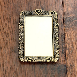 May include: A rectangular picture frame with a metallic gold finish and a dark, antiqued patina. The border features a raised, repeating relief pattern of sculpted roses, leaves, and small heart shapes surrounding a plain, off-white center.