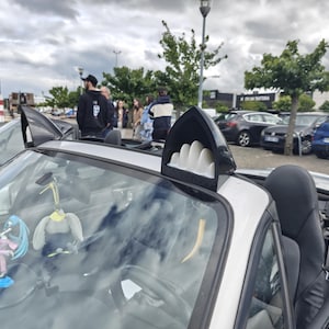 Pode incluir: Um carro conversível prateado com um spoiler de barbatana de tubarão personalizado. O interior do carro é preto e o para-brisa tem objetos decorativos pendurados. O fundo mostra um estacionamento com outros carros e pessoas.