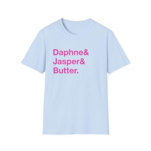 May include: A light blue t-shirt with the words "Daphne & Jasper & Butter." printed in pink. The shirt has short sleeves and a crew neck. The text is centered on the front of the shirt.