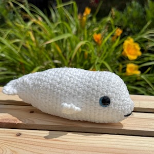 May include: A handmade white crochet beluga whale plush toy with small fins and a smiling mouth. The whale has blue eyes and is resting on a wooden surface. The background features green foliage and yellow flowers.