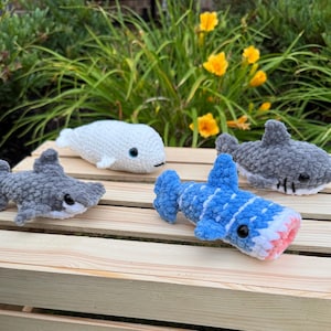 May include: Crocheted plush toys of a whale and sharks in various colors. The toys include a white whale, two gray sharks, and a blue shark with a pink and white mouth. The toys are placed on a wooden crate.