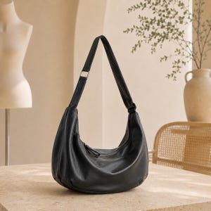May include: A black hobo bag with a smooth, curved silhouette and a single shoulder strap. The bag features a top zipper closure with a matching black pull tab, a soft, rounded base, and a silver-toned metal buckle on the strap for adjustment.